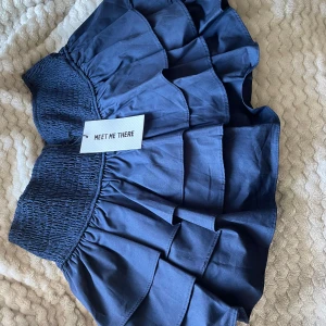 Marinblå volangkjol Meet Me There - Supersöt marinblå kortkjol från Meet Me There med bred smock i midjan och flera lager volanger. Kjolen har innershorts för extra komfort. Perfekt för dig som gillar en elegant och trendig stil. Nypris ca 400kr