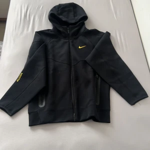 Svart NOCTA x Nike hoodie med dragkedja - Svart hoodie från Nike NOCTA med hel dragkedja och huva. Gula detaljer som Nike-logga på bröstet och NOCTA-tryck på ärmen. Fickor med dragkedja och diskreta broderade initialer bak. Perfekt för en sportig och stilren look. Storlek /s