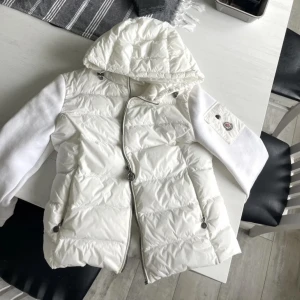 Vit pufferjacka från Moncler med huva - Säljer en snygg vit pufferjacka från Moncler med avtagbar huva och dragkedja framtill. Jackan har långa ärmar i mjukt material och två fickor med knappar. Perfekt för kyliga dagar och riktigt stilren look. Passar fint även på vår/sommae 