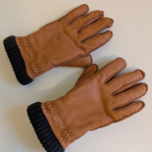 Hestra Deerskin Primaloft Rib handskar, storlek 7 - Snygga bruna skinnhandskar från Hestra med svart ribbad mudd. Handskarna är varma och skinnet mjukt och är perfekta för kyliga dagar. Märket är diskret ingraverat på ovansidan. Använda några gånger under förra vintern.