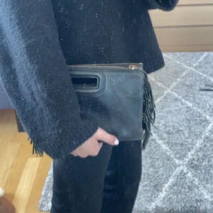 Svart maje kuvertväska med fransar - Säljer en svart maje kuvertväska i skinn med dekorativa fransar på sidorna och dragkedja upptill. Väskan har ett praktiskt handtag och stilren design, perfekt för att bära det viktigaste.
