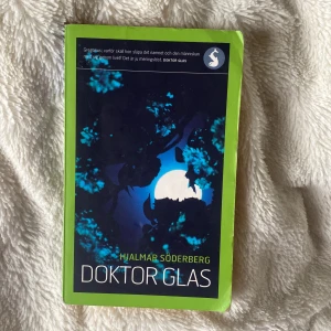 Doktor Glas - Säljer romanen 'Doktor Glas' av Hjalmar Söderberg. Pocketbok med grön ram och ett omslag i mörkblått med silhuett av träd och måne. En svensk klassiker om moral, kärlek och existentiella frågor. Perfekt för dig som gillar psykologiska romaner.