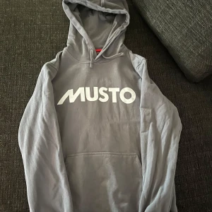 Grå hoodie från Musto - Säljer en grå hoodie från Musto med stor vit logga på bröstet. Tröjan har huva med dragsko och en stor magficka framtill. Perfekt för en avslappnad stil. Ny pris 700 ( pris kan diskuteras)