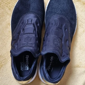 Svarta Reebok Classic sneakers med gulddetalj - Säljer ett par svarta Reebok Classic sneakers med snygg guldfärgad detalj på hälen och vit sula med svarta stänk. Ovandelen är i syntet och mocka, utan snörning för en clean look. Perfekta för dig som vill sticka ut med stilrena detaljer.