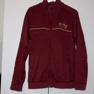 Vinröd zip-jacka från BOSS - Säljer en vinröd zip-jacka från BOSS med guldfärgade detaljer och logga på bröstet. Jackan har hög krage, dragkedja och ribbade muddar. Snygga kontrastsömmar och BOSS-tryck på insidan av kragen.  De finns två små hål som syns på tredje bilden annars i bra skick! 