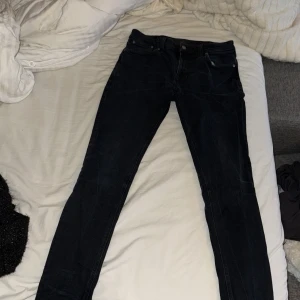 Svarta jeansbyxor från Nudie Jeans - Säljer ett par svarta jeans från Nudie Jeans med klassisk femficksdesign och raka ben. Jeansen har silverfärgade nitar och knapp, samt Nudie-logga på bakfickan och läderpatch i midjan.