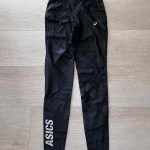 Svarta träningstights från Asics - Svarta tights från Asics med logga längs ena benet och liten logotyp på höften. Perfekta för träning med stretchigt material och smal passform. Elastisk midja för extra komfort.