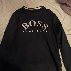 Svart sweatshirt från Hugo Boss - Säljer en svart sweatshirt från Hugo Boss med stort vitt BOSS-tryck på bröstet. Tröjan har rund halsringning och långa ärmar. Perfekt för en stilren och avslappnad look. Tröjan passar suveränt till storlek M också.