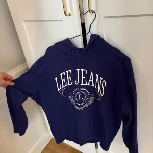 Blå hoodie från Lee Jeans - Säljer en blå hoodie från Lee Jeans med vit logga framtill. Tröjan har huva med dragsko och långa ärmar. Perfekt för en avslappnad stil.