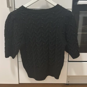 Svart stickad tröja med pärlor - Säljer en svart stickad tröja med vackert mönster och små pärlor över hela tröjan. Tröjan har halvlånga ärmar och bred ribbad mudd nedtill. Perfekt för dig som vill ha något extra till din outfit.