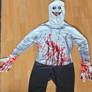Blodig Morphsuit - Säljer en vit och svart morphsuit med blodstänk och läskigt ansikte på huvan. Dräkten har dragkedja på baksidan och tryckta blodiga detaljer på armar och händer. Perfekt för halloween eller maskerad! Finns en liten fläck på baksida huvud men inget som är synligt, det fanns en peruk till men den är tyvärr borttappad. 
