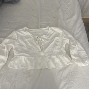 Vit croppad blus med knyt - Säljer en söt vit croppad blus med långa ärmar och knytning framtill. Blusen har volang nertill och en lätt v-ringning. Perfekt till vår och sommar för en fräsch look.