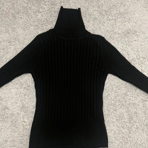 Svart ribbad polotröja - Säljer en svart ribbad polotröja med lång ärm. Tröjan är tight i modellen och har en hög polokrage som ger en stilren look. Perfekt att matcha med jeans eller kjol för en snygg outfit. Den har ingen storleksmärkning, men passar dig som vanligtvis har XXS, XS eller S. Perfekt för kyligare dagar – stilren, enkel och skön att bära!