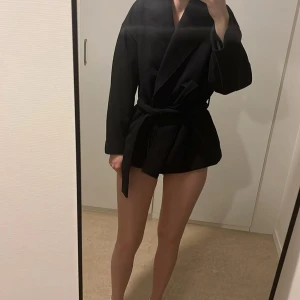 Svart kappa Ginatricot  - Säljer en svart kappa och knytbälte i midjan. Kappan har en oversized passform och är långärmad, perfekt för en stilren look. Passar till många olika outfits och är enkel att matcha.