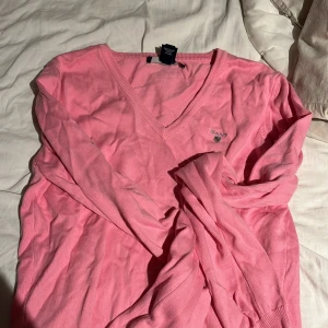 Rosa v-ringad tröja från GANT - Säljer en rosa v-ringad tröja från GANT med broderad logga på bröstet. Tröjan har lång ärm och är tillverkad i mjuk bomull. Perfekt för dig som gillar klassisk och stilren design.