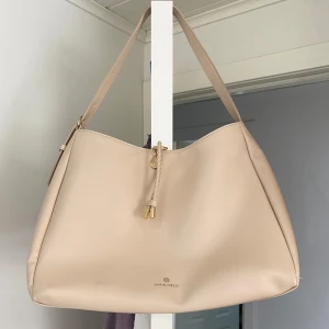 Beige axelväska från Anna Field - Snygg beige axelväska från Anna Field med justerbart axelband och dekorativ flätad snodd framtill. Väskan har guldfärgade detaljer och en stilren, enkel design som passar till många outfits.