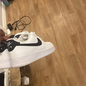 Nike Air Force 1 vita sneakers - Klassiska Nike Air Force 1 sneakers i vitt med svart swoosh. Skorna har snörning, Tillverkade i syntetmaterial med tydlig Nike-logga på hälen och tungan. Perfekta för en stilren och sportig look. Använda 1 gång 