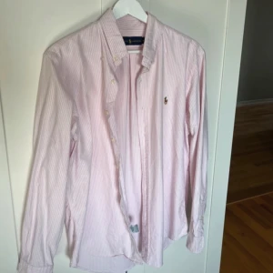 Rosa och vit randig skjorta från Ralph Lauren - Klassisk långärmad skjorta från Ralph Lauren i rosa och vitt med tunna ränder. Skjortan har knappar framtill och på kragen samt en broderad logga på bröstet. Perfekt för en stilren och tidlös look. 