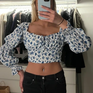 Blommig croppad blus med puffärmar - Superfin croppad blus med blått blommönster och långa puffärmar. Toppen har en söt knytning framtill och smockad midja som ger en snygg passform. Perfekt för vår och sommar! storlek s!