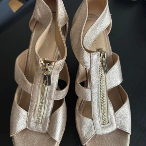 Guldiga wedge-sandaler från Michael Kors - Säljer ett par snygga guldiga sandaler från Michael Kors med kilklack och dragkedja framtill. Skorna har öppen tå och flätad sula i repimitation, perfekt för sommaren. Supersnygga till både jeans och klänning!
