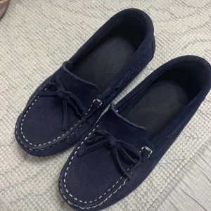 Mörkblå mockaloafers med snördetalj - Säljer ett par mörkblå loafers i mocka med dekorativ snörning framtill och kontrastsömmar. Skorna har en platt sula med gummidetaljer undertill för extra grepp. Perfekta för en stilren och avslappnad look. För barn.
