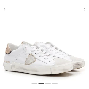 Philippe Model skor - Snygga vita sneakers med guldmetallic detalj på hälen och dekorativt hjärta i beige på sidan. Skorna har snörning och en något smutsad look på sulan för en trendig känsla. Perfekta för dig som vill sticka ut med stilrena men unika detaljer. Box och påse med kommer om man vill. Nypris 4000kr