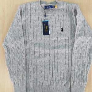 Ljusgrå stickad tröja från Polo Ralph Lauren - Säljer en klassisk ljusgrå stickad tröja från Polo Ralph Lauren med kabelstickat mönster och broderad logga på bröstet. Tröjan har rund halsringning och långa ärmar. Perfekt för en stilren och tidlös look.