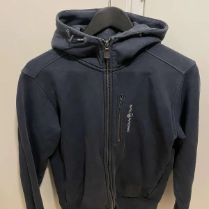 Mörkblå hoodiejacka från Sail Racing - Mörkblå hoodiejacka från Sail Racing med dragkedja framtill och bröstficka med logga.