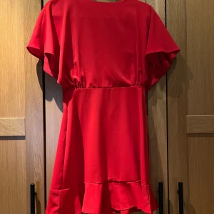 Röd klänning med volang - Säljer en röd klänning med korta vida ärmar och volangdetalj nertill. Klänningen har en markerad midja och är tillverkad i ett slätt, lätt material. Perfekt för dig som vill sticka ut med färg!