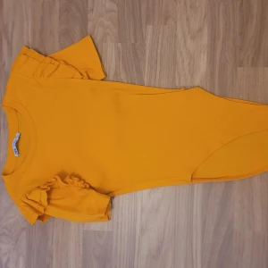 Gul ribbad body med volangärmar - Säljer en kort gul body med ribbad struktur och volangdetaljer på ärmarna. Korta ärmar och en figurnära passform som framhäver siluetten. Perfekt för dig som vill sticka ut med färg!