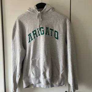 Grå hoodie från Arigato - Säljer min gråa hoodie från Arigato i herrstorlek M (EU 48–50). Den har krympt något i tvätten, så den passar bäst om du ligger mellan S och M. Fortfarande skön och i bra kvalitet!  Det finns två små hål vid magfickan – inget som stör funktionen, men bra att känna till.