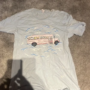 Ljusblå K-12 Tour t-shirt från Melanie Martinez - Ljusblå t-shirt med tryck av en buss fylld med tecknade djur och texten 'K-12 Tour' på framsidan. Baksidan har turnélistan och fler illustrationer. Perfekt för fans av Melanie Martinez och hennes K-12 era.