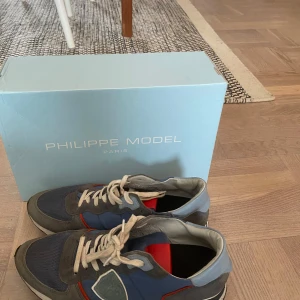 Blå Philippe Model sneakers med grå och röda detaljer - Snygga sneakers från Philippe Model i blått med grå och röda detaljer. Skorna har vita snören, rund tå och platt sula. Materialmix med inslag av mocka och syntet. Perfekta för en trendig och avslappnad stil.