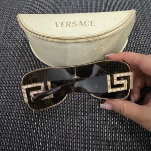 Svarta solglasögon från Versace med gulddetaljer - Snygga svarta solglasögon från Versace med stora, fyrkantiga glas och exklusiva guldfärgade detaljer i geometriskt mönster på sidorna. Kommer med originalfodral. Perfekt accessoar för att sticka ut.