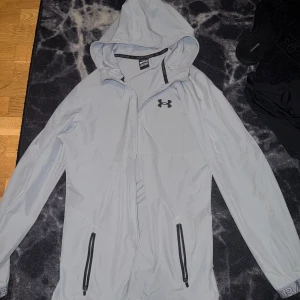 Ljusgrå vindjacka från Under Armour - Säljer en ljusgrå vindjacka från Under Armour med huva och svarta dragkedjor. Jackan har två fickor framtill och en diskret logga på bröstet. Perfekt för utomhusaktiviteter och träning.