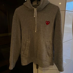 Grå hoodie från Comme des Garçons Play - Tjena🙌🏻 säljer en Grå hoodie från Comme des Garçons Play med klassisk röd hjärtlogga på bröstet. Tröjan har huva med dragsko och känguruficka framtill. Perfekt för en avslappnad och stilren look. Tröjan är äkta
