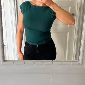 Mörkgrön topp med öppen rygg - Säljer en mörkgrön tight topp med korta ärmar och snyggt öppen rygg. Toppen har en enkel, stilren design och är tillverkad i ett stretchigt material som sitter skönt mot kroppen.
