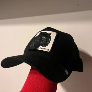 Svart keps med pantermärke från Goorin Bros - Svart keps från Goorin Bros med ett broderat märke framtill som föreställer en panter och texten 'Panther'. Kepsen har böjd skärm och justerbar baksida för perfekt passform.