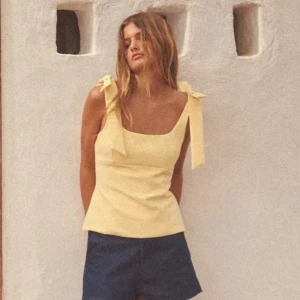 Gul linnetopp med rosetter på axlarna - Söt ljusgul linnetopp med breda axelband som knyts i stora rosetter. Toppen har en fyrkantig urringning och är figurnära i modellen. Perfekt för varma dagar och ger en fräsch sommarkänsla.