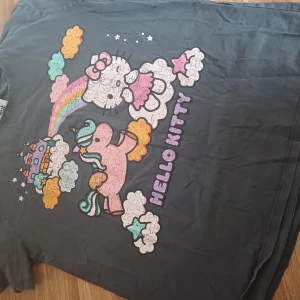 Hello Kitty t-shirt från Zara - Grå t-shirt från Zara med stort Hello Kitty-tryck framtill. Motivet har färgglada moln, regnbåge, enhörning och slott i pastellfärger. T-shirten har rund hals och korta ärmar. Perfekt för dig som älskar söta och lekfulla prints!
