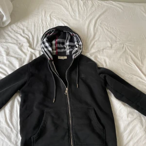 Svart hoodiejacka från Burberry med rutigt foder - Svart hoodiejacka från Burberry med dragkedja och klassiskt rutigt foder i huva och insida. Jackan har fickor framtill och justerbar huva med snören. Perfekt för en stilren och avslappnad look. Size M passar S 