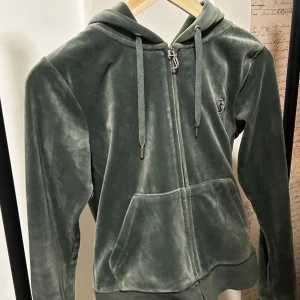 Grön velour hoodie från Juicy Couture - Mjuk och skön grön hoodie och byxa i velour från Juicy Couture. Koftan har dragkedja framtill, huva och stora fickor. Byxor med snören och silver detaljer långt ut. Även på baksidan på rumpan finns två fickor med knappar som även har silver detaljer. Perfekt för en avslappnad och trendig look.