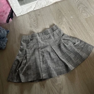 Grå rutig kjol med veck - Säljer en grå och svart rutig kjol med klassiskt rutmönster och bred linning. Kjolen har fina veck som ger en snygg form och är i kort modell. Perfekt för dig som gillar preppy stil.
