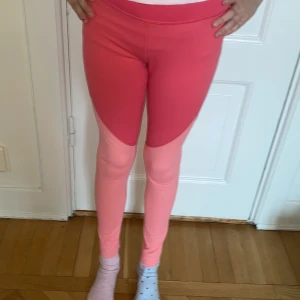 Tvåfärgade träningstights från SOC - Säljer ett par snygga träningstights från SOC i två nyanser av rosa. Tightsen har en tight passform och är gjorda i ett stretchigt syntetmaterial som andas. Perfekta för träning eller sportaktiviteter.