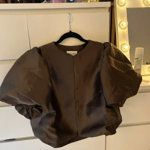 Brun cleo blus by Malina - Säljer en brun blus från by Malina i modellen Cleo. Blusen har stora puffärmar och rund halsringning. Perfekt för dig som vill sticka ut med en trendig och elegant topp.