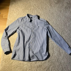 Blårandig skjorta från Polo Ralph Lauren - Säljer en klassisk blårandig skjorta från Polo Ralph Lauren i slim fit. Barnstorlek 20 motsvara i vuxenstorlek XS/S.Skjortan har långa ärmar, knappar framtill och den ikoniska broderade loggan på bröstet. Perfekt för dig som gillar stilrena och tidlösa plagg.