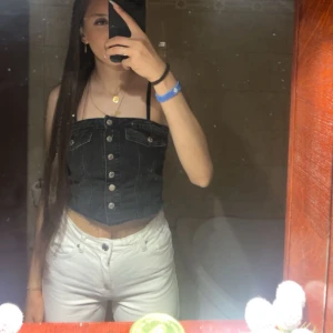 Svart jeans korsett-topp med knappar - Säljer en svart croppad korsett-topp i jeansmaterial med silvriga knappar framtill och råa kanter upptill. Toppen har dekorativa fickor och smock baktill för bättre passform. Perfekt för att lyfta vilken outfit som helst!