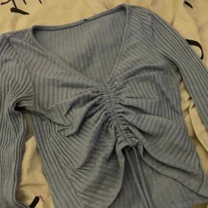 Ljusblå ribbad topp med knyt - Trendig ljusblå långärmad topp med ribbat material och v-ringning. Toppen har snygg knyt- och rynkdetalj framtill som ger en cool look. Perfekt att styla med jeans eller kjol.
