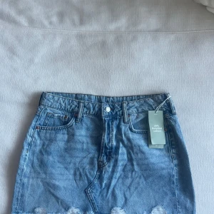 Blå jeanskjol från A Denim - Säljer en klassisk blå jeanskjol från A Denim med råa kanter nertill och femficksmodell. Kjolen har en normal passform och stängs med knapp framtill. Perfekt att matcha med en enkel topp eller hoodie.