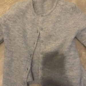 Ljusgrå kofta från Gina Tricot - Mysig ljusgrå kofta från Gina Tricot med rund halsringning och knäppning framtill. Koftan har långa ärmar och är tillverkad i ett mjukt, stickat material. Perfekt att slänga på sig när det är kyligt ute!